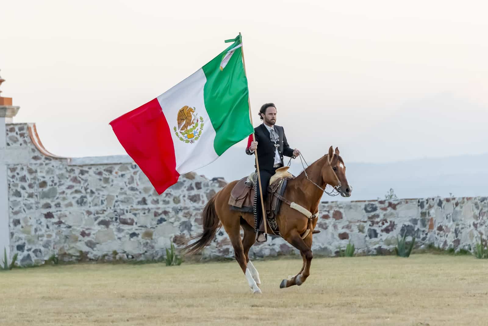 33097136 a very handsome mexican charro poses in front of a hacienda in the mexican countryside while holding the mexican flag